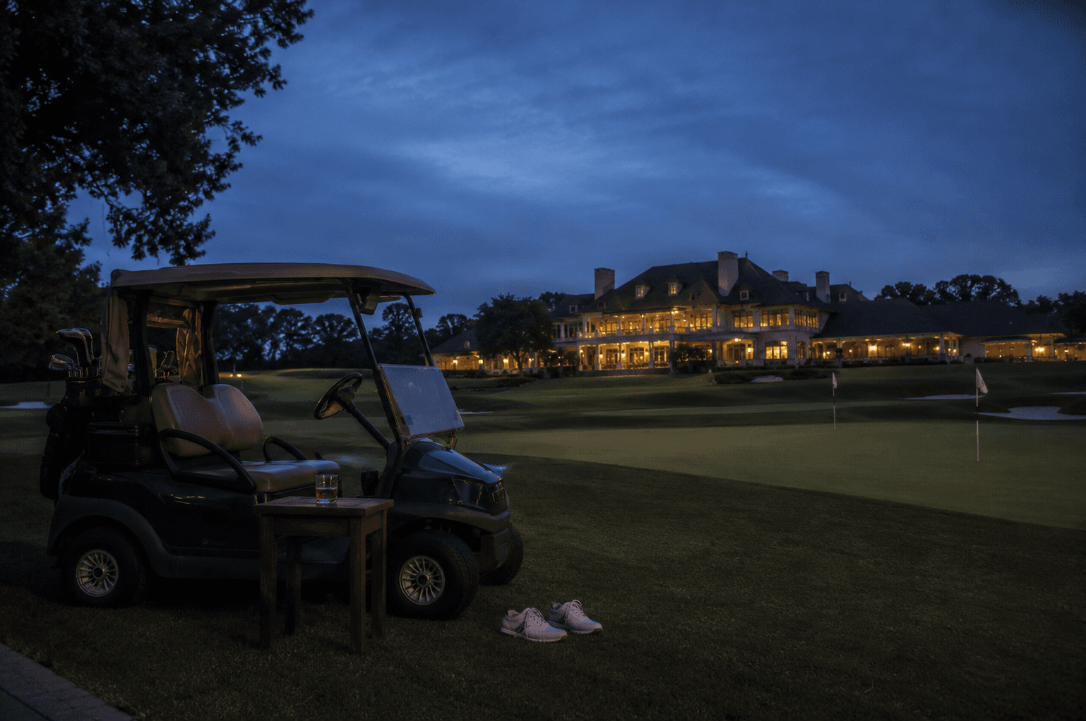 A manicured golf course at dusk with an empty clubhouse glowing in the distance, a single abandoned cart at the edge of the green